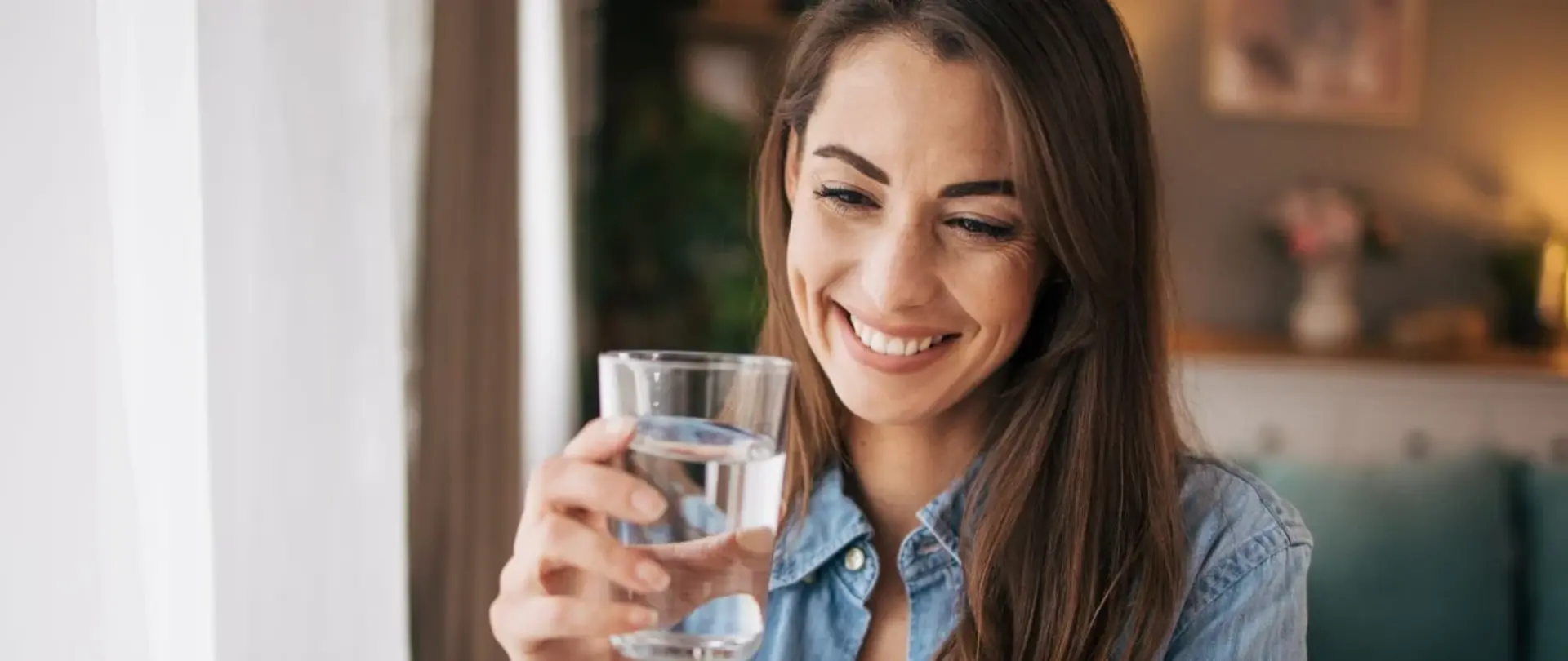 Eine Frau steht am Fenster und hält ein Glas Wasser in der Hand. Sie trägt ein Jeans-Hemd und eine weiße Bluse