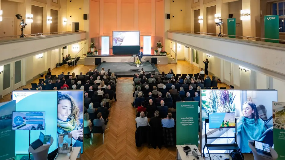 Man sieht das Publikum von hinten mit Fokus auf Banner und Aufsteller hinter dem Publikum.