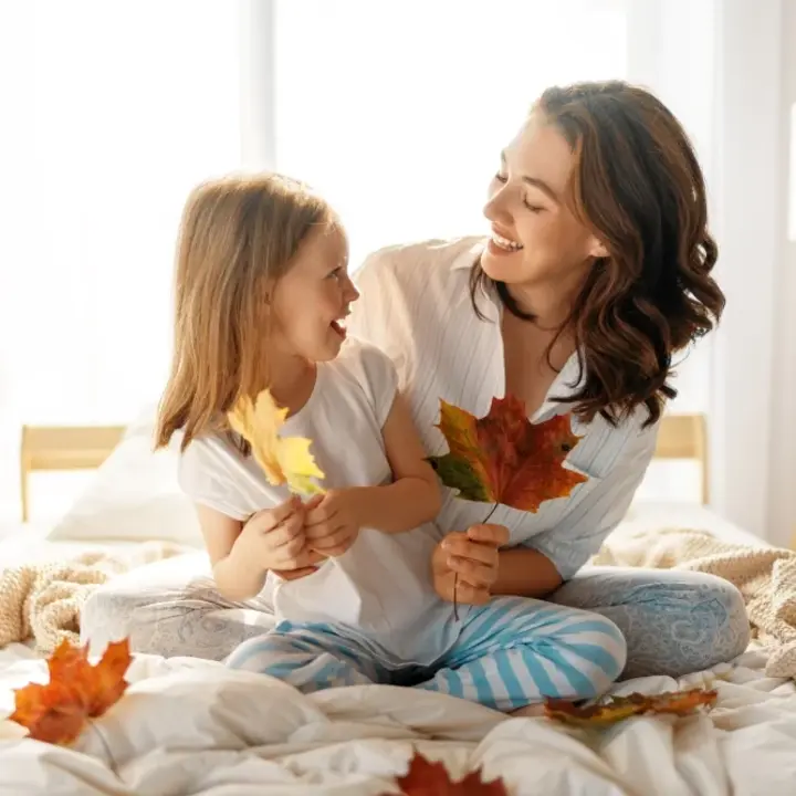 Eine Mutter sitzt mit ihrer kleinen Tochter auf dem Bett. Sie haben herbstliche Ahornblätter in den Händen und lächeln sich an. Es ist ein sehr helles und gemütlich anmutendes Foto