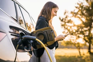 Eine Person steht neben einem ladenden Elektroauto und hält ein Smartphone in der Hand.