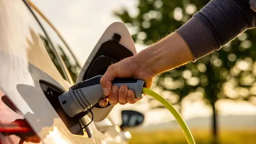Eine Hand steckt ein Ladekabel in den Ladeanschluss eines weißen Elektroautos im Freien.
