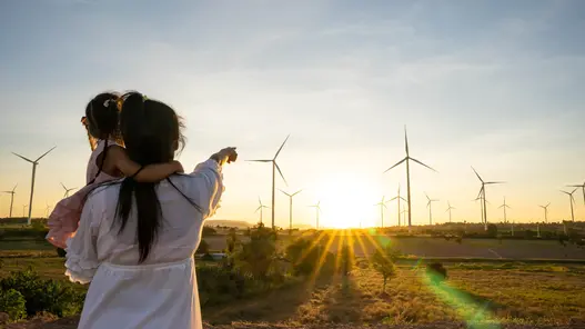 Eine Mutter hat ihre Tochter auf dem Arm und zeigt auf einen Windpark im Hintergrund beim Sonnenuntergang.
