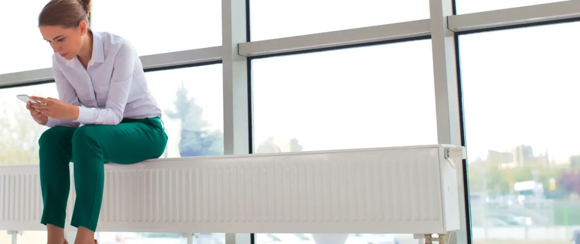 Eine Frau sitzt auf einer Heizung vor einer großen Fensterfront und ist nach vorne gelehnt. Sie schaut auf ihr Handy.