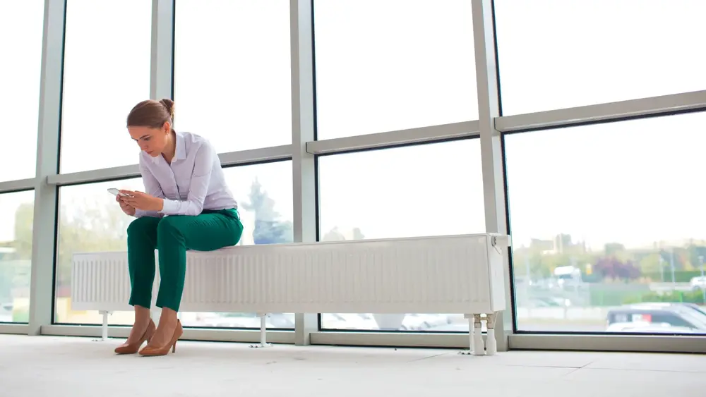 Eine Frau sitzt auf einer Heizung vor einer großen Fensterfront und ist nach vorne gelehnt. Sie schaut auf ihr Handy.