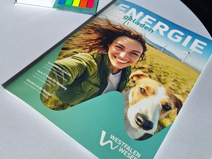 Der Titel der zweiten Ausgabe des Kundenmagazins ENERGIEgeladen zeigt eine Frau mit ihren zwei Hunden auf einer Wieso vor Windkraftanlagen.