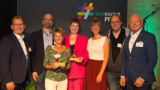 Platz 3: New Norm Paderborn Gruppenfoto vor einer Fotowand von Vereinsmitgliedern von New Norm Paderborn mit dem 3. Hauptpreis des WWKulturpreis26.