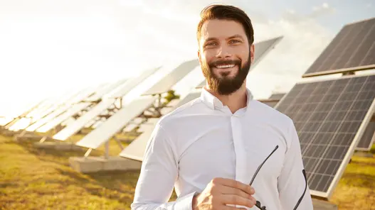 Ein lächelnder Mann in Anzug und mit Brille in der Hand steht auf einer Wiese vor vielen Photovoltaikmodulen. 