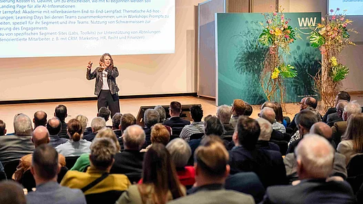 Anna Kopp referiert vor Publikum Man sieht das Publikum von hinten mit Fokus auf die Referentin Anna Kopp, die auf der Bühne steht und einen Vortrag hält.