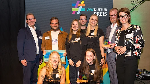 Platz 2: Festival Burgbeben STRNBG Dörentrup Gruppenfoto vor einer Fotowand von Vereinsmitgliedern des Festival Burgbeben STRNBG mit dem 2. Hauptpreis des WWKulturpreis26.