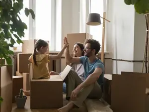 Eine dreiköpfige Familie sitzt im Wohnzimmer ihrer neuen Wohnung umringt von Kartons. Der Vater und das Kind klatschen ein