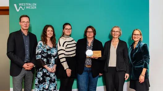 Sechs Personen stehen vor einem großen Westfalen Weser Hintergrund. Die Dame in der Mitte hält die Auszeichnung in ihrer linken Hand.