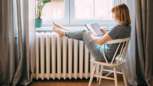 Eine Frau sitzt auf einem Stuhl am Fenster, liest ein Buch und hat die Füße auf der Heizung abgelegt