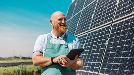 Ein Unternehmer steht vor einer PV-Freiflächenanlage und schaut lächelnd auf sein Tablet..