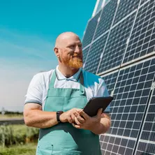 Ein Unternehmer steht vor einer PV-Freiflächenanlage und schaut lächelnd auf sein Tablet..