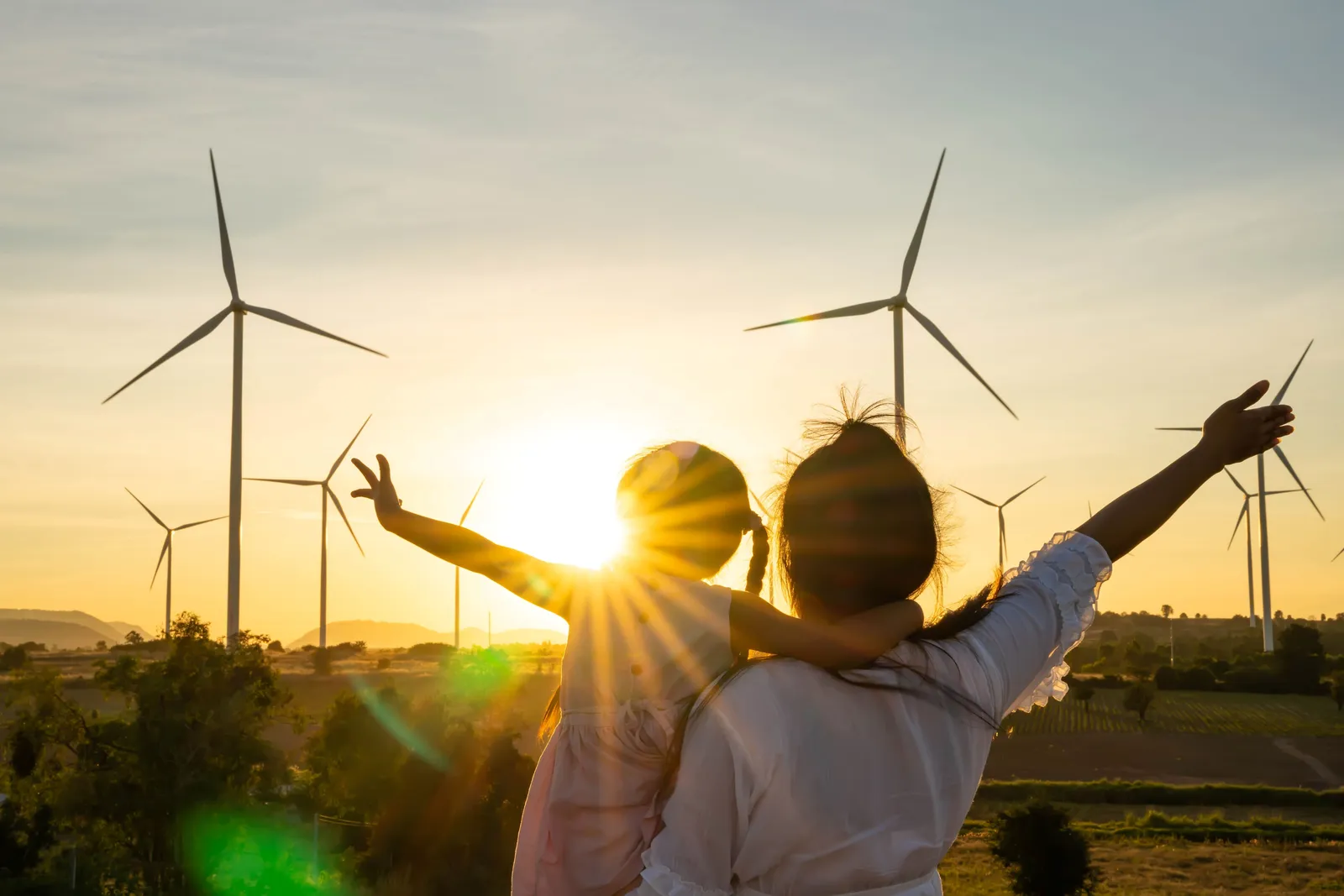 Eine Person hält ein Kind im Arm, beide blicken auf Windkraftanlagen bei Sonnenuntergang.