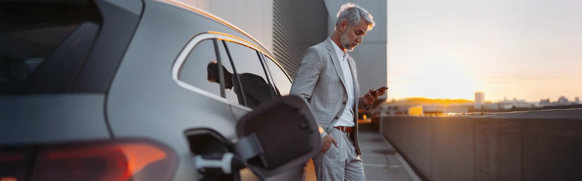 Eine Person im Anzug lädt ein Elektroauto und nutzt ein Smartphone. auf einem Parkplatz bei Sonnenuntergang.