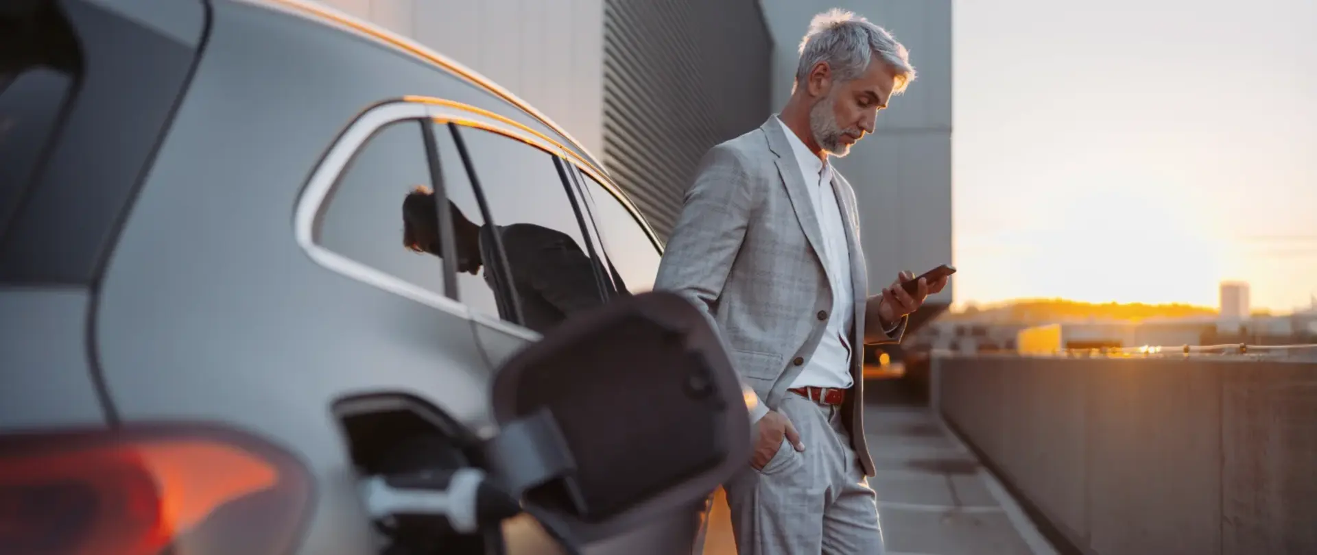 Eine Person im Anzug lädt ein Elektroauto und nutzt ein Smartphone. auf einem Parkplatz bei Sonnenuntergang.