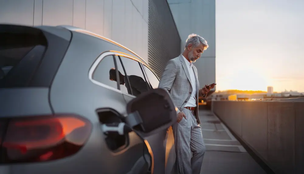 Eine Person im Anzug lädt ein Elektroauto und nutzt ein Smartphone. auf einem Parkplatz bei Sonnenuntergang.