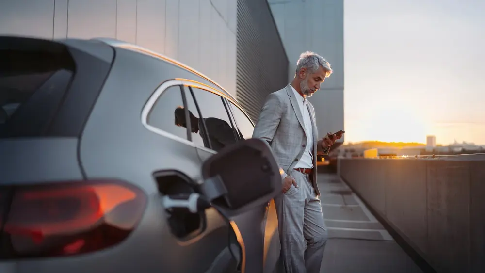 Eine Person im Anzug lädt ein Elektroauto und nutzt ein Smartphone. auf einem Parkplatz bei Sonnenuntergang.