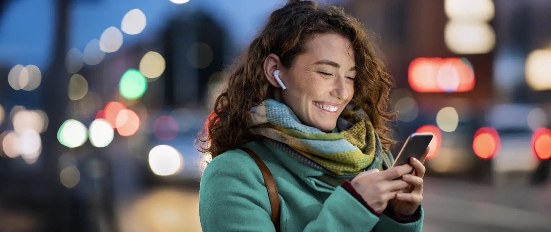 Eine Person mit Smartphone und Kopfhörern steht abends in einer beleuchteten Stadtumgebung mit verschwommenem Straßenhintergrund.