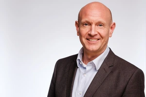 Ein freundlich lächelnder Mann im dunklen Anzug vor einem hellgrauen Hintergrund.