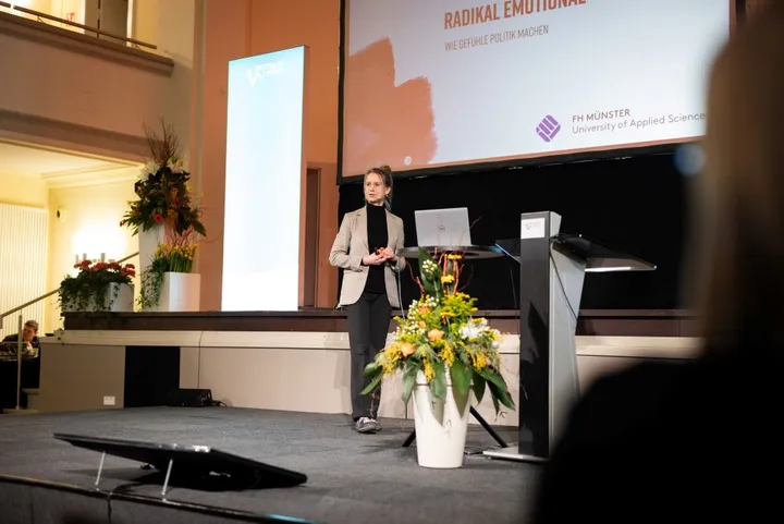 Westfalen Weser Forum Rede Man sieht eine junge Frau auf einer Bühne eine Rede halten mit einem großen Bildschirm im Hintergrund.