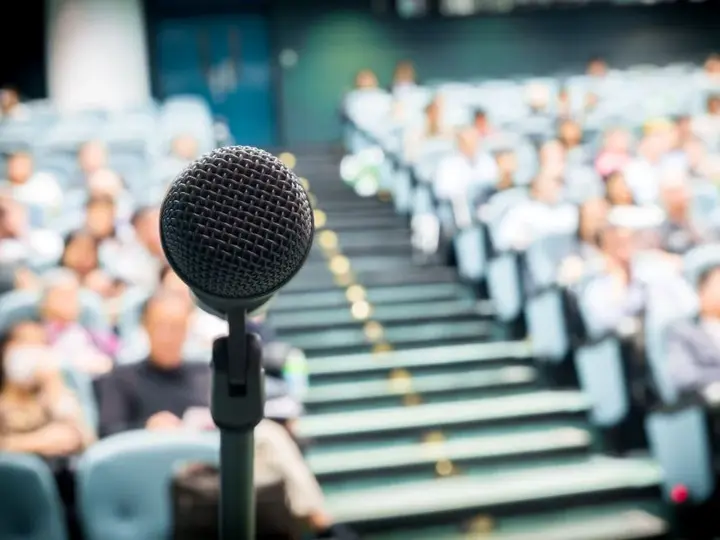 Man sieht ein Mikrofon im Fokus und im Hintergrund das Publikum unscharf